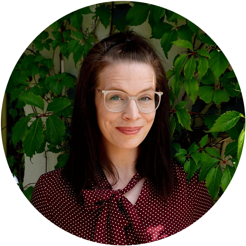 Mitarbeiterin-von-Pat.nrw Mitarbeiterin von Pt.nrw mit braunen schulterlangen haaren und einer Brille. Steht vor grünen Pflanzen