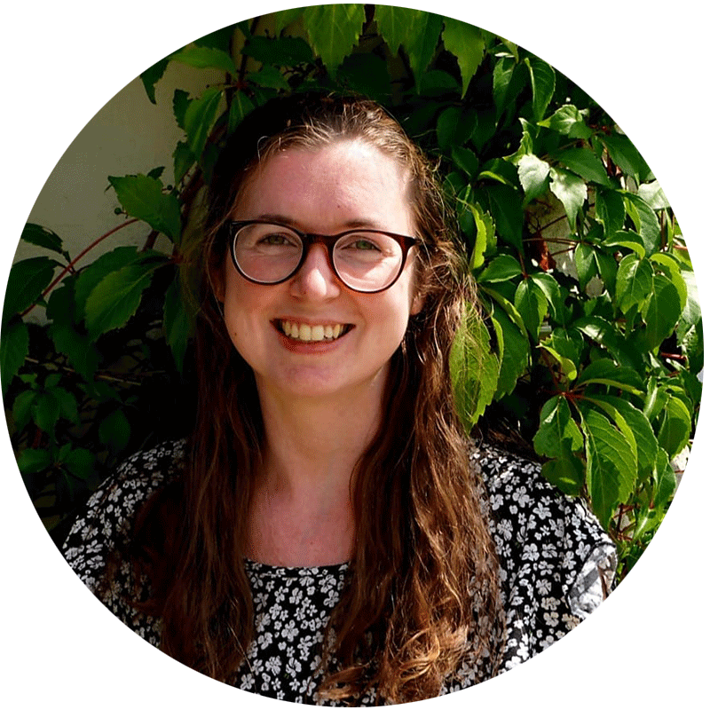 Mitarpeiterin-von-pat.nrw Mitarbeiterin von Pat.nrw. mit dunkelblonden lockigen haaren und einer Brille, lächelt in die Kamera und steht vor grünen Pflanzen